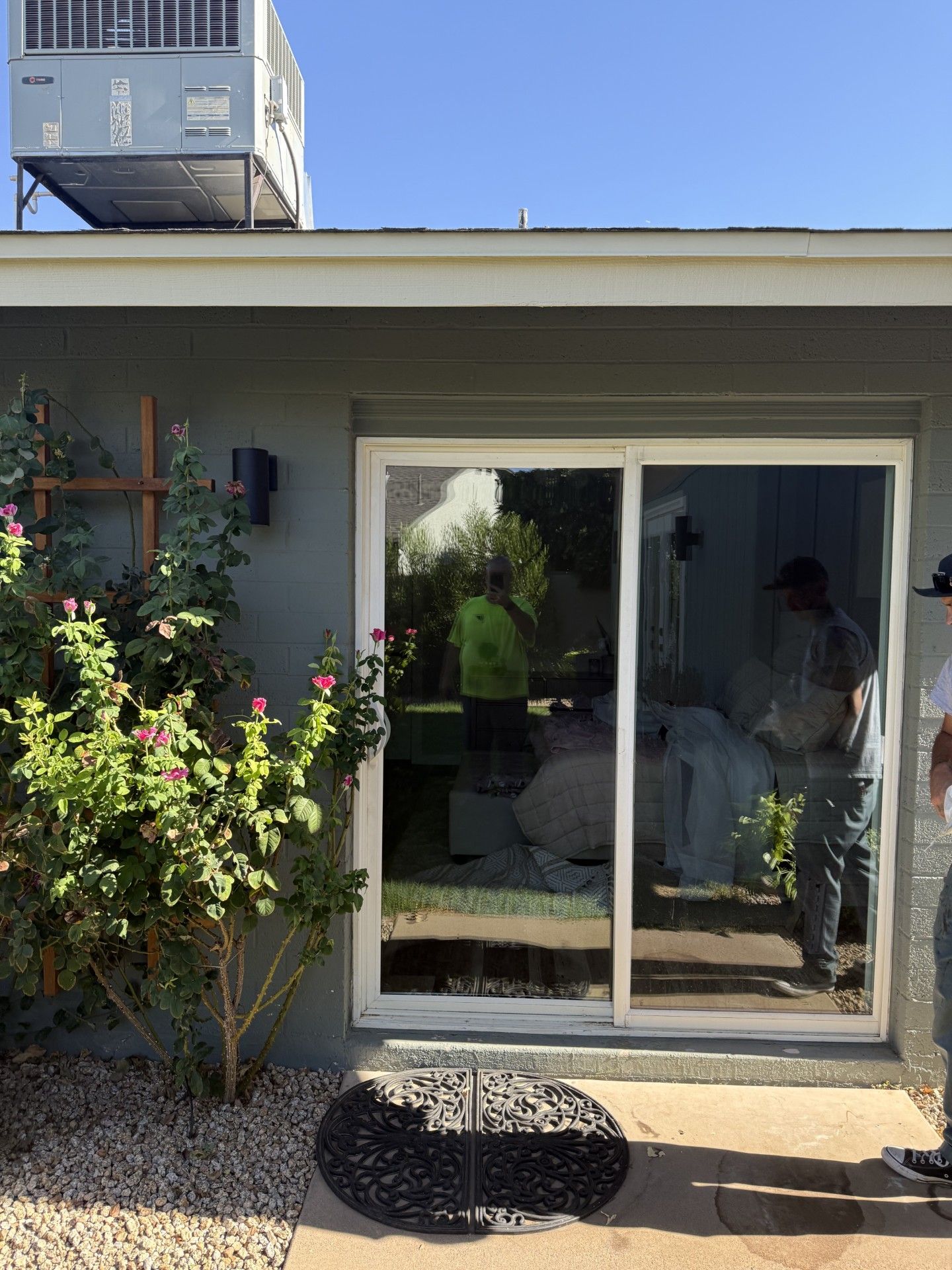 Sliding glass door reflects workers. Exterior view with AC unit, plants, and green house paint.