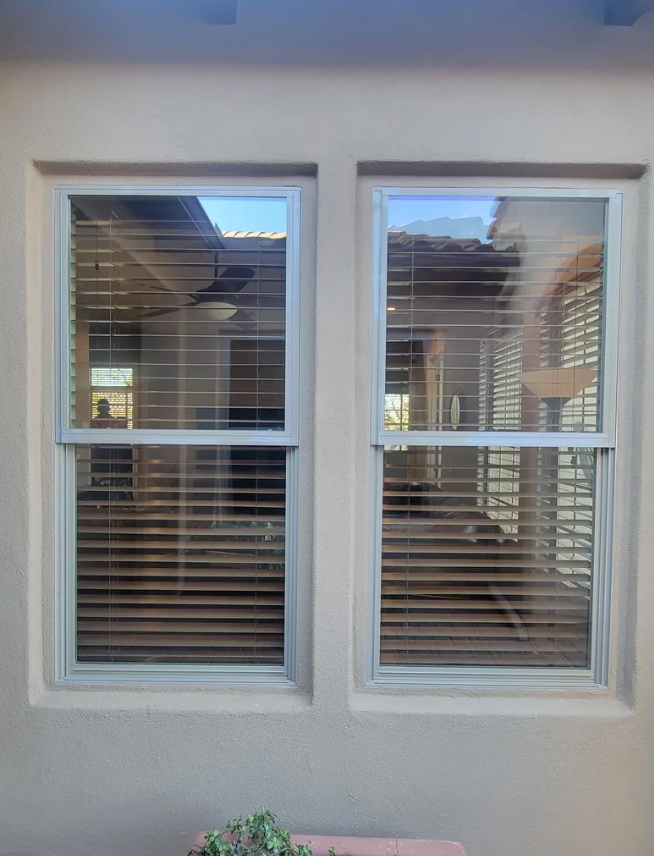 Two white-framed windows with closed blinds reflecting a patio setting, set in a tan stucco wall.
