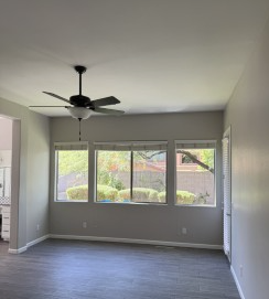 Empty living room with gray walls, large windows, ceiling fan, and dark flooring.