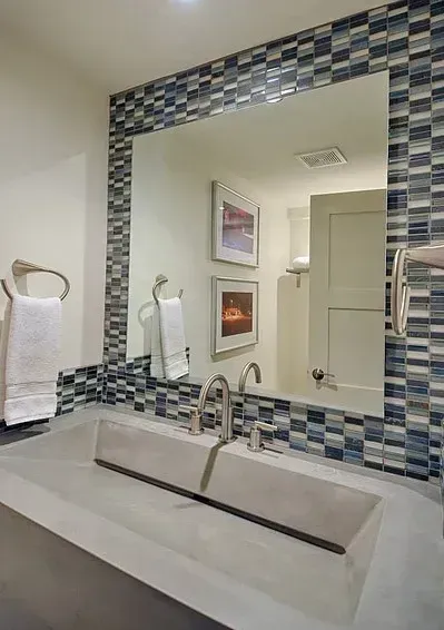 Bathroom with a large mirror framed by blue and gray tiles, concrete sink, and white towels.