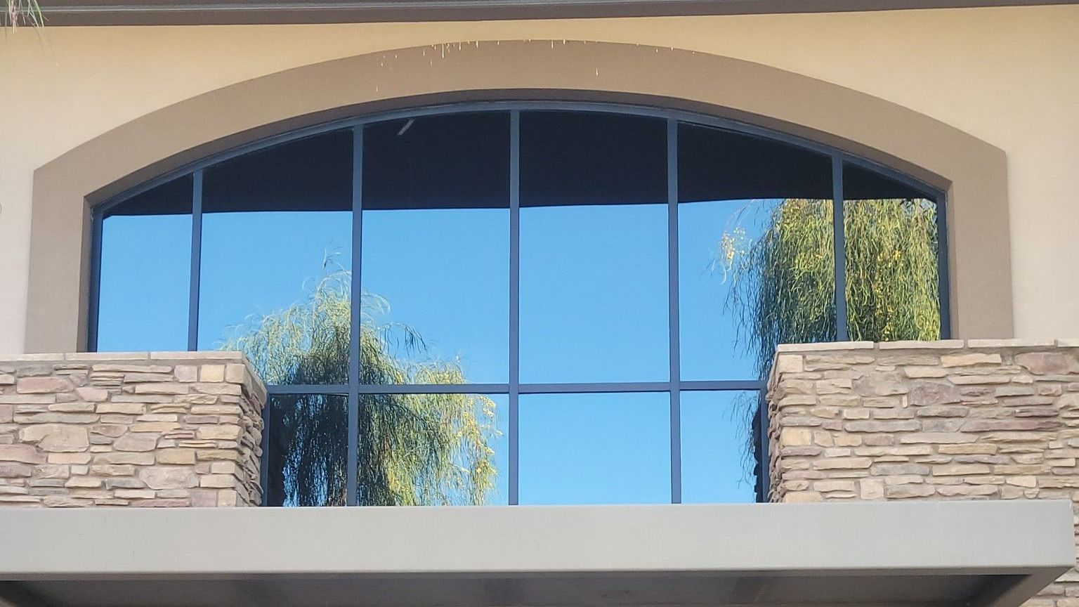 Large arched window reflecting a blue sky and trees, framed by a stone and stucco exterior.