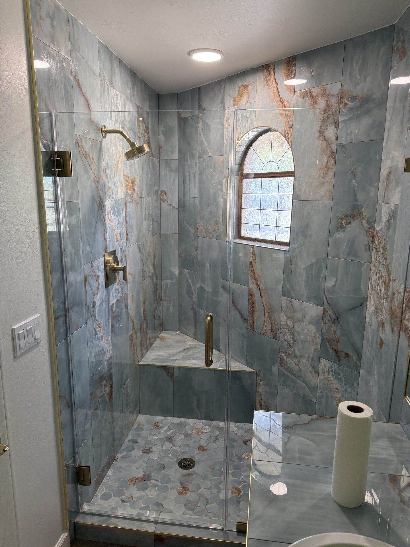 Blue marble tiled shower with glass doors, brass fixtures, and an arched window.