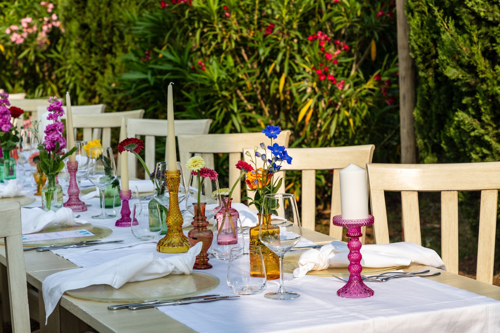 Tavolo da pranzo all'aperto apparecchiato con drappeggi bianchi, candelabri colorati e fiori in un giardino.