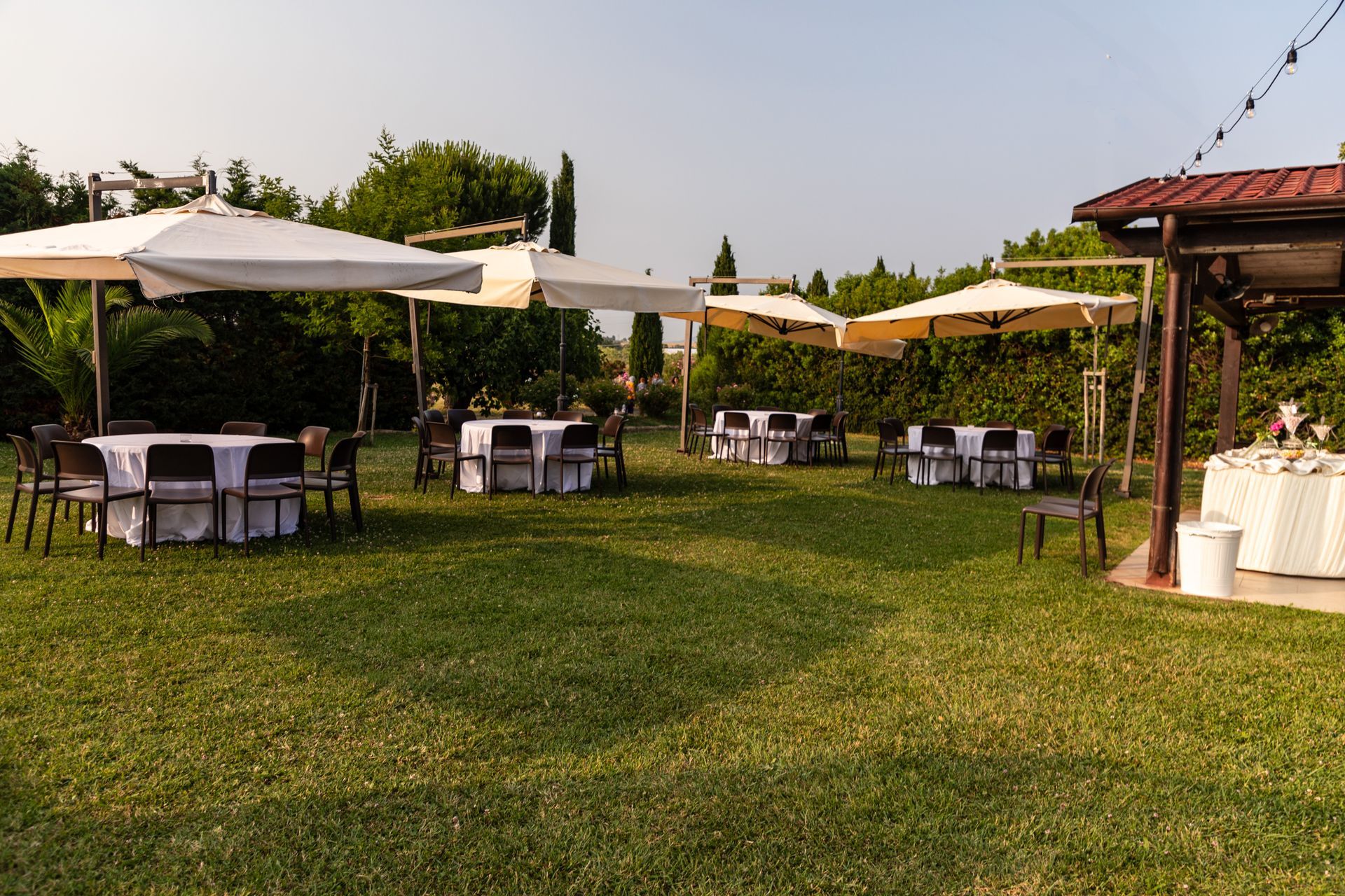 Allestimento per eventi all'aperto in giardino con ombrelloni bianchi, tavoli e luci decorative su un prato verde.
