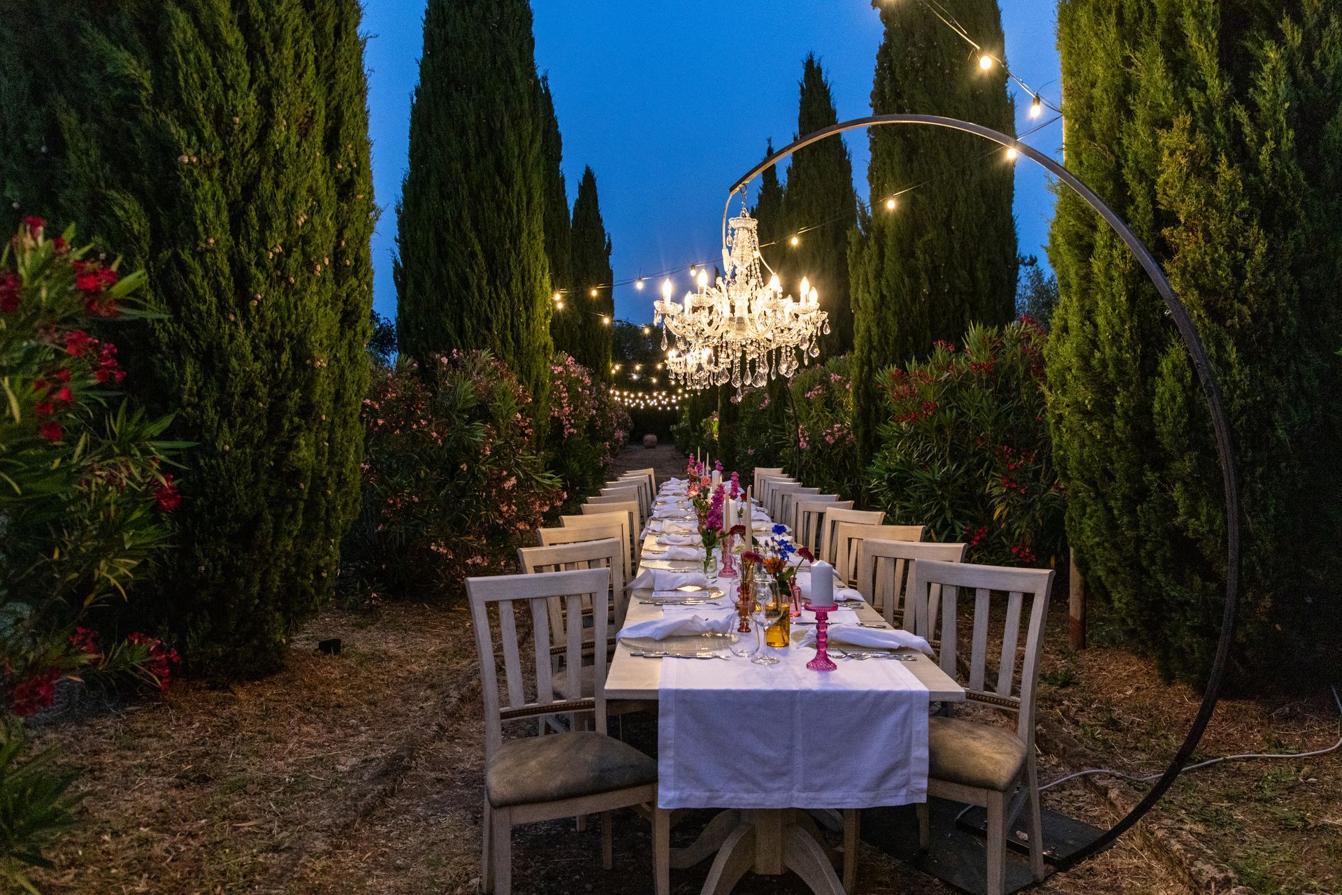 Un lungo tavolo da banchetto sotto i lampadari in un giardino fiancheggiato da alti cipressi al crepuscolo