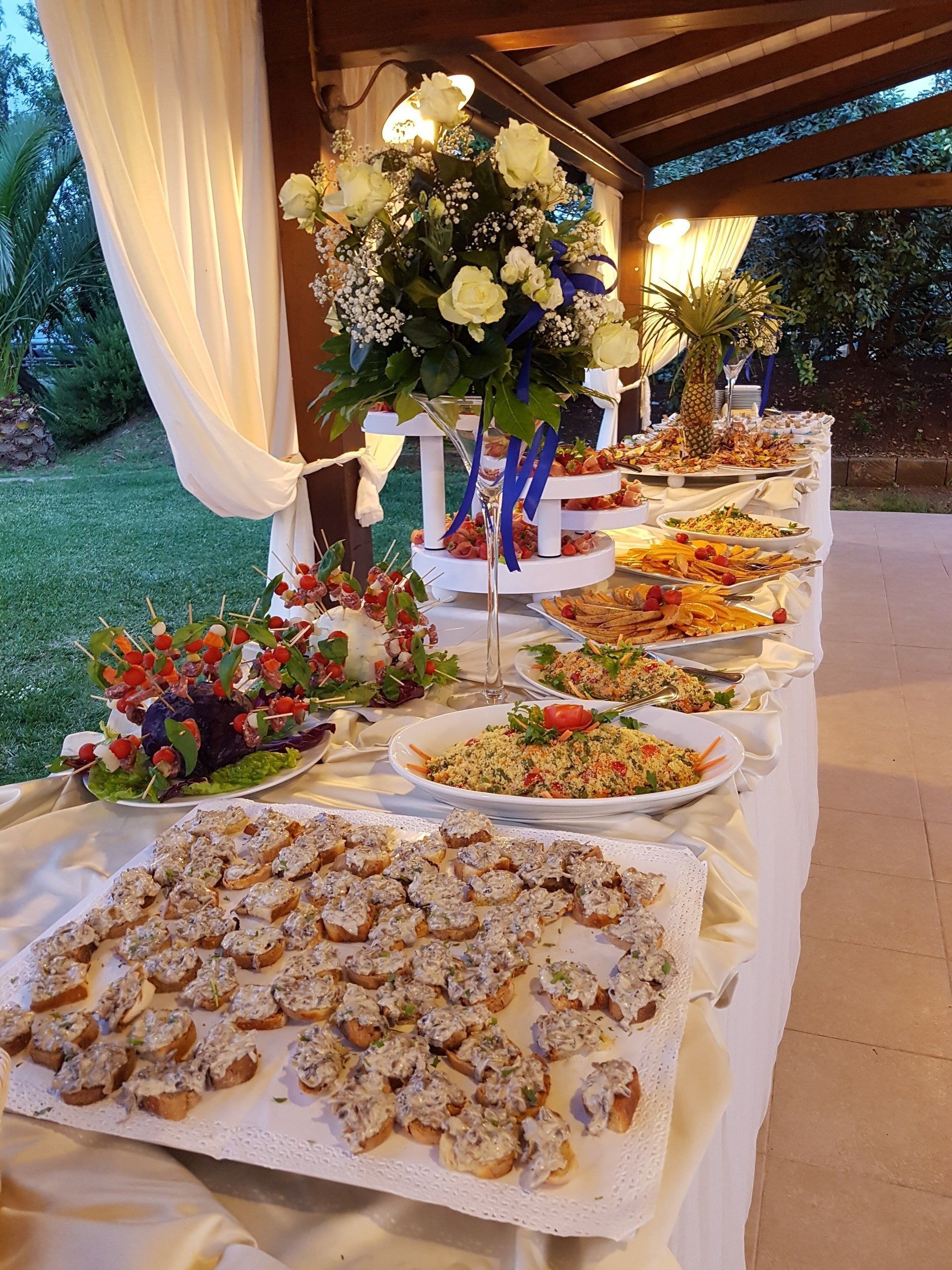 Tavolo da buffet con assortimento di stuzzichini e un grande centrotavola floreale per un evento all'aperto.