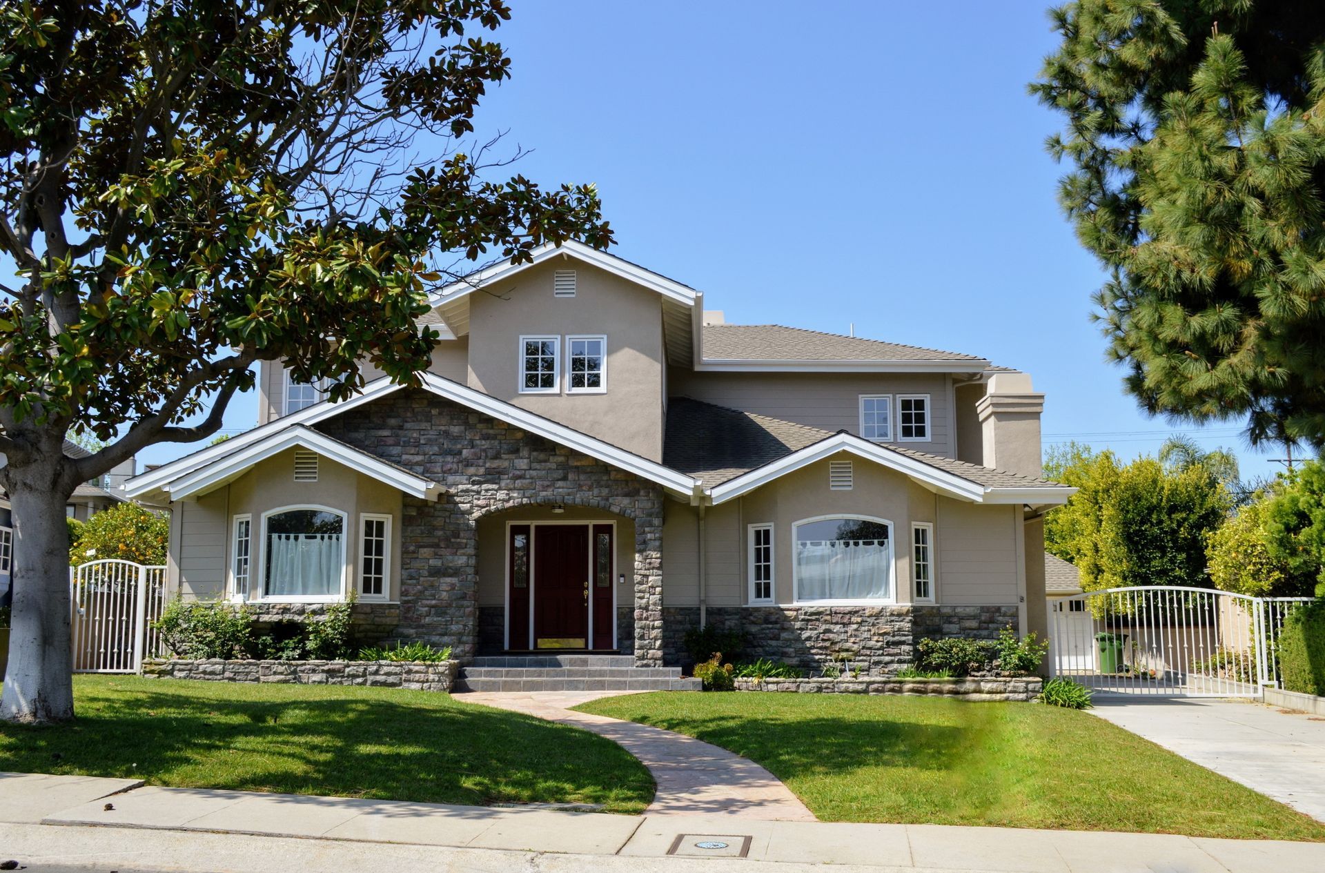 Two-story tan house with stone accents, arched windows, and a red door under a sunny blue sky.