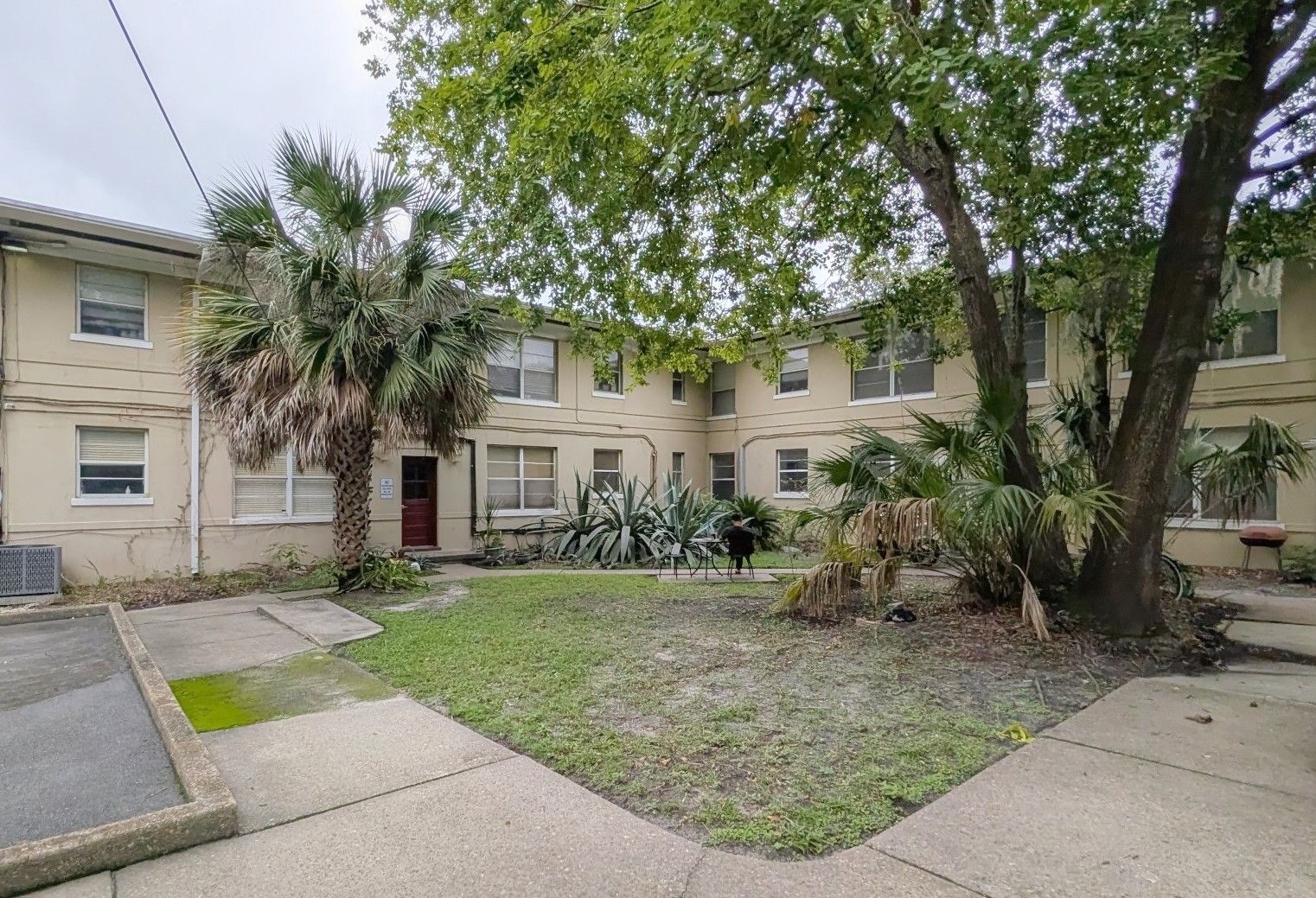 Exterior view of a two-story apartment building with a small yard. Beige walls, windows, palm trees, and overcast sky.