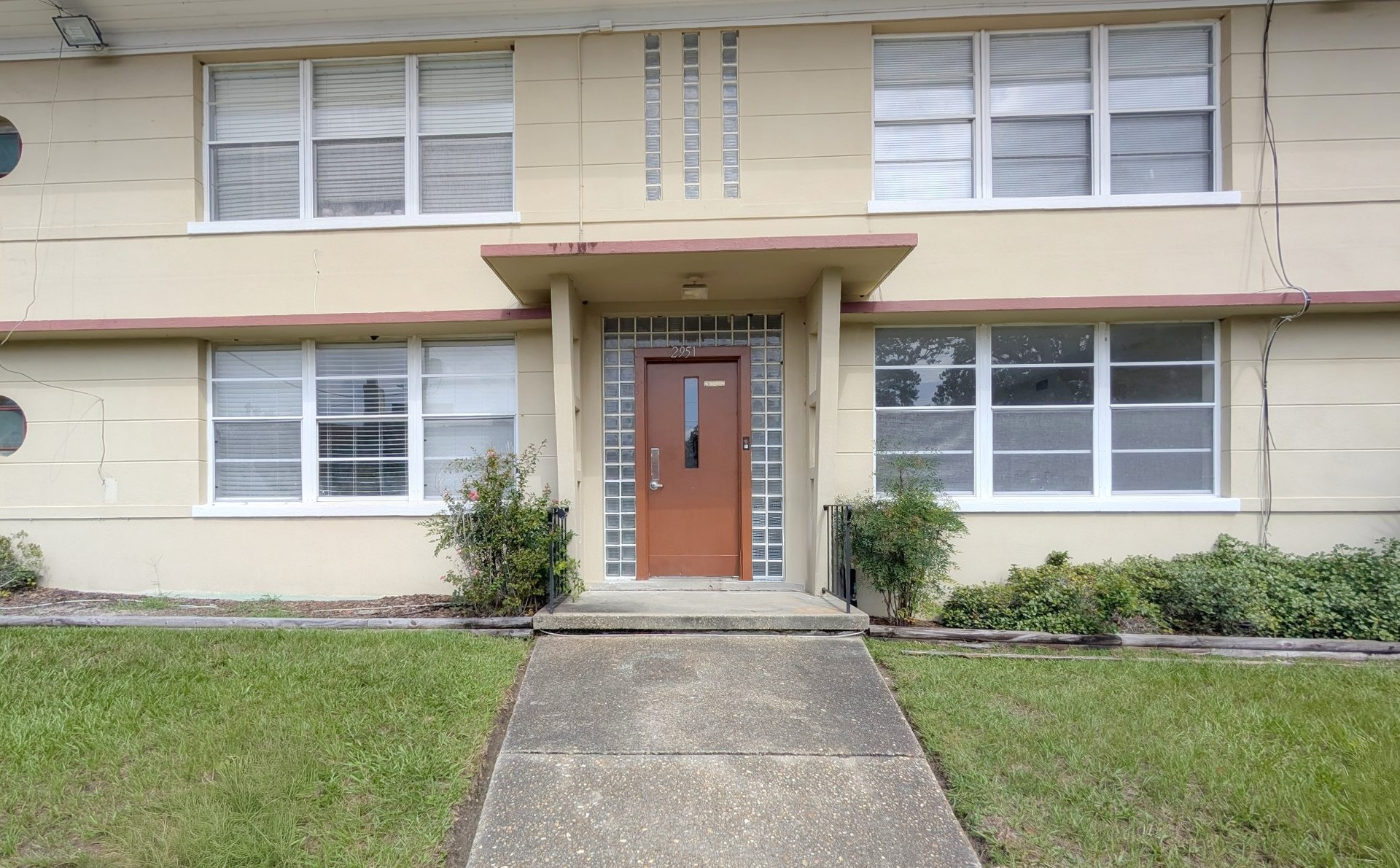 Two-story beige apartment building with white-framed windows, a brown door, and a concrete walkway.