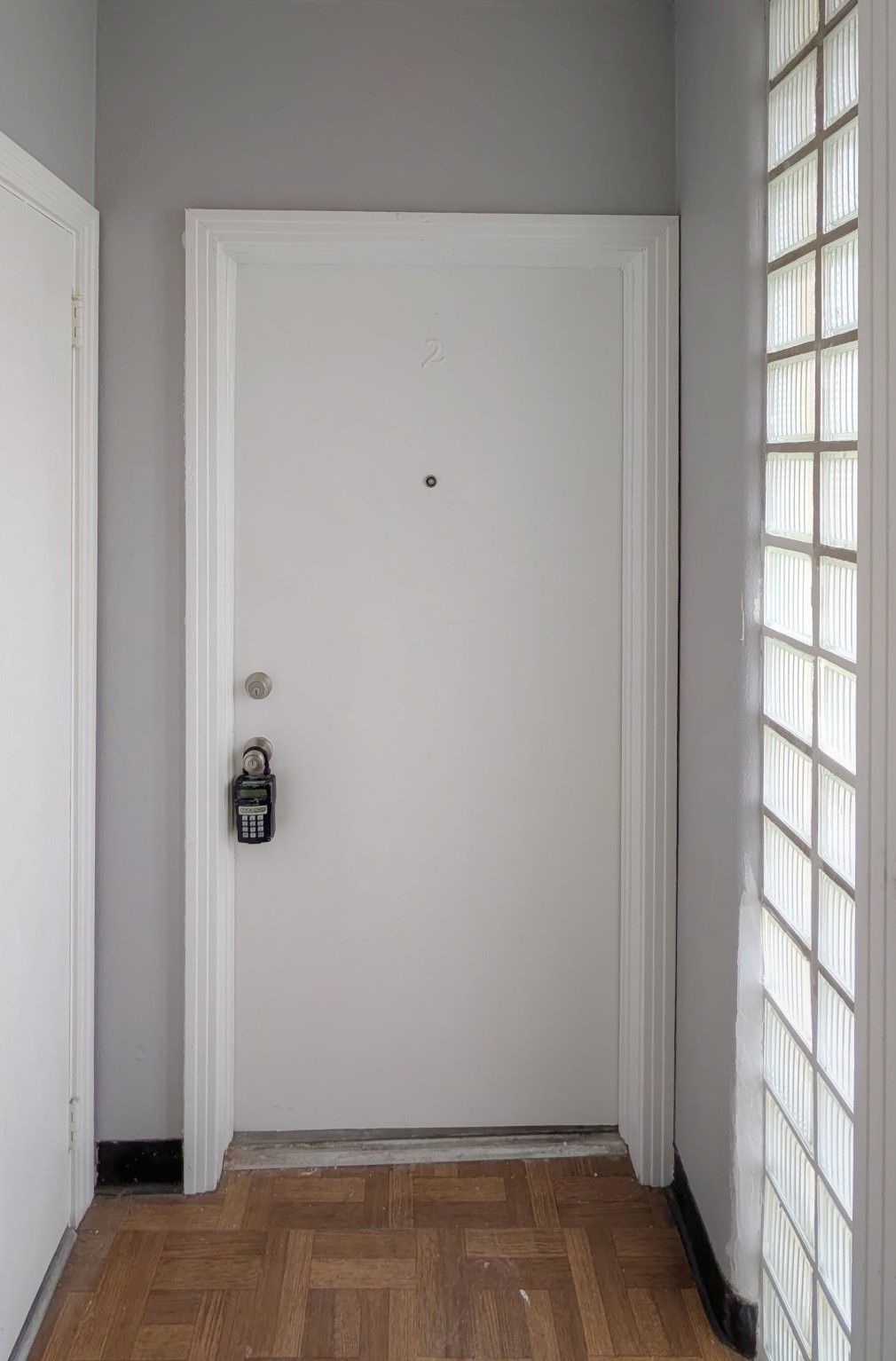 White door with a peephole and lock, in a gray hallway with a glass block window.