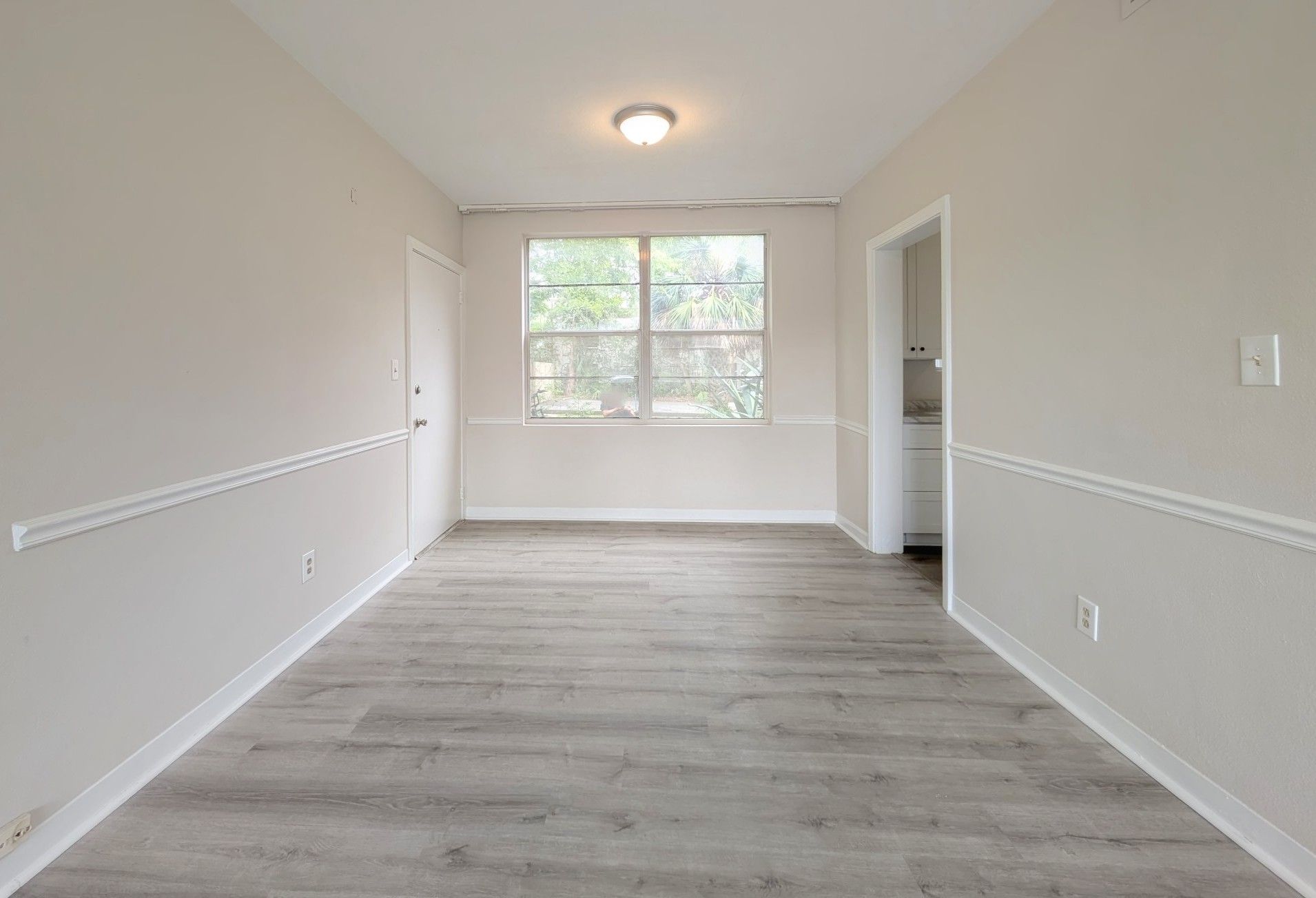 Empty dining room with gray wood-look flooring, light walls, a window, and doorway to the kitchen.
