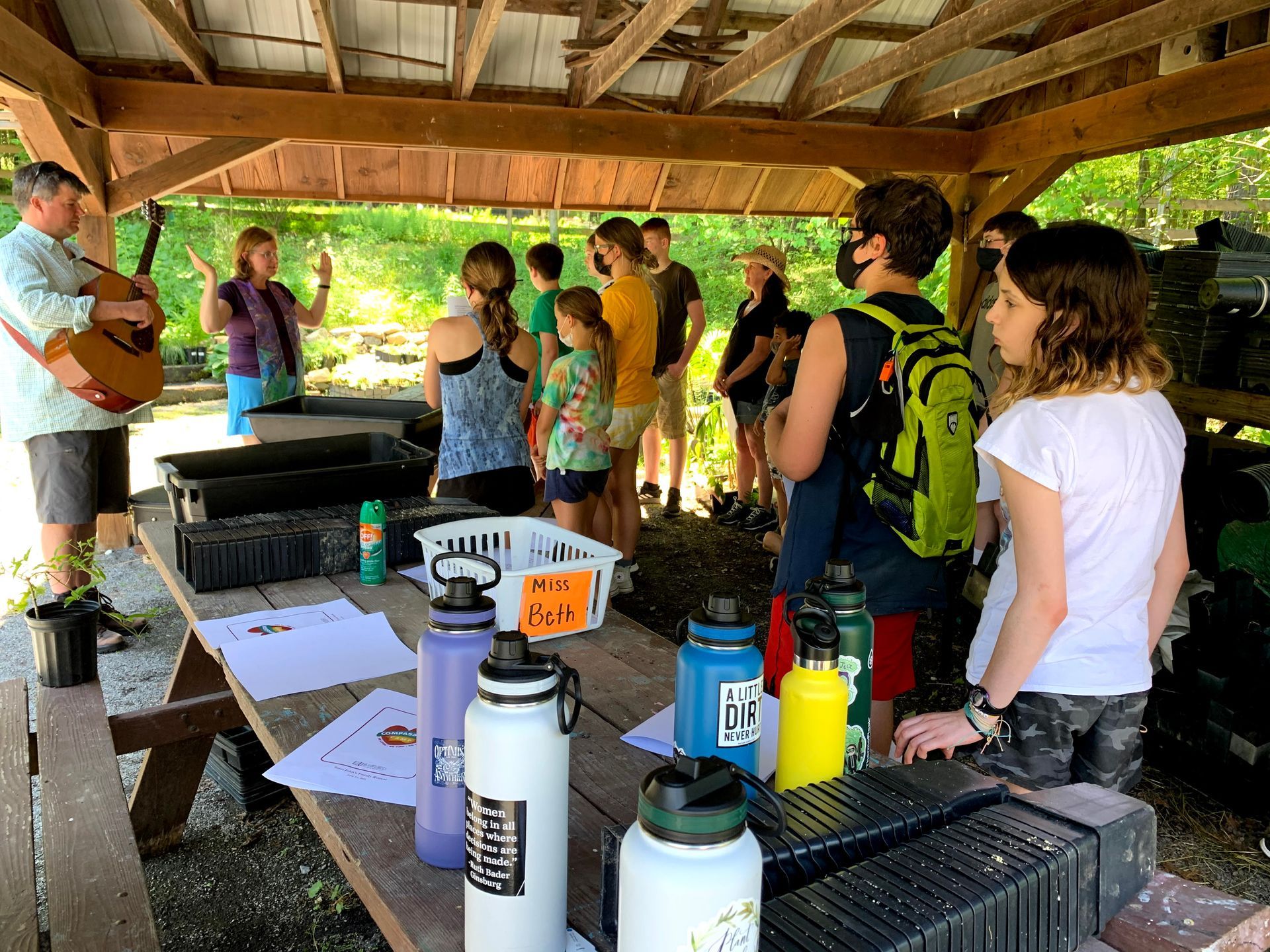 A group of people standing around a picnic table with water bottles and a man playing a guitar