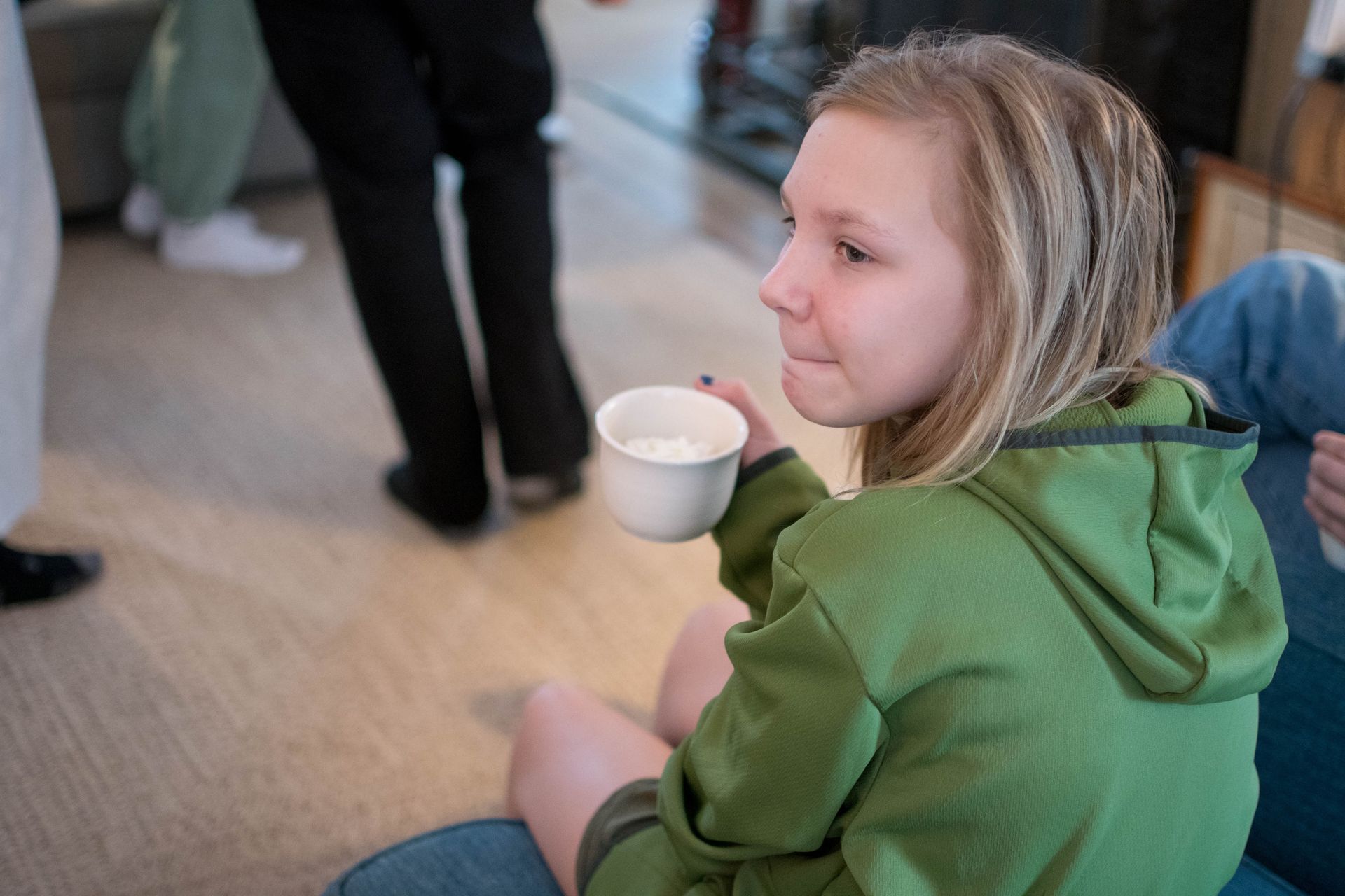 A girl in a green jacket is drinking from a white cup