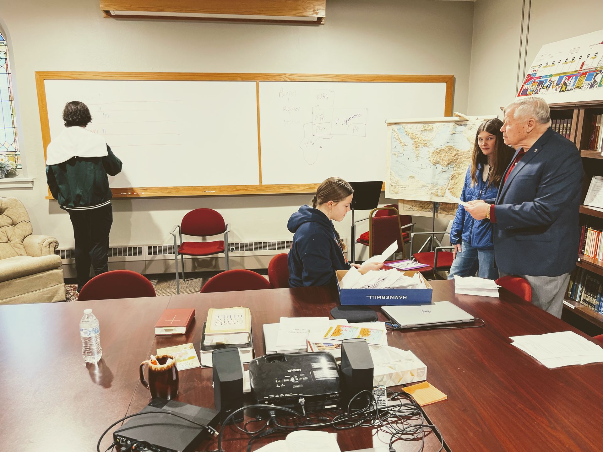 A group of people are gathered around a table in front of a white board