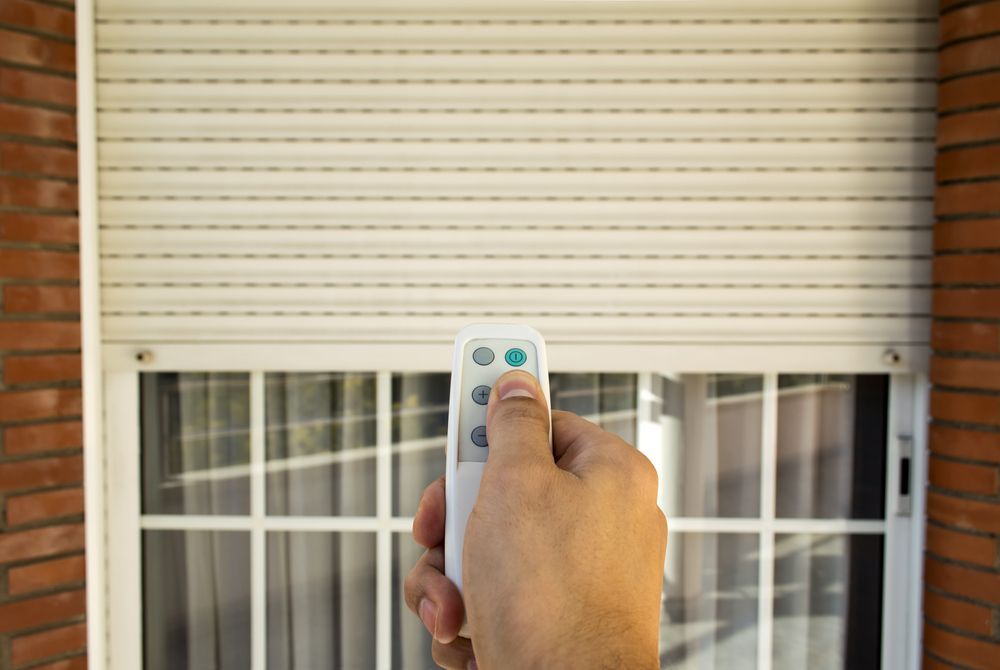 Hand Holding a Remote Control, Operating a White Shutter Over a Window — Industry Fire Services in Charters Towers, QLD