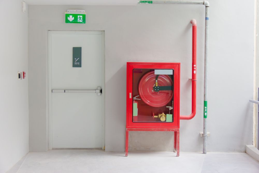 Red Fire Hose Cabinet Next to a White Door With an Exit Sign, in a Hallway — Industry Fire Services in Weipa, QLD