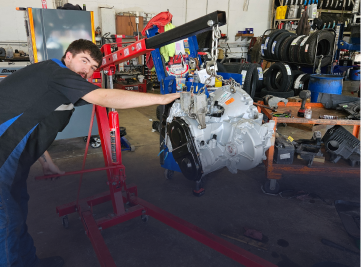 Mechanic points to engine suspended by a red hoist in a garage. | Z-1 Automotive, Inc.