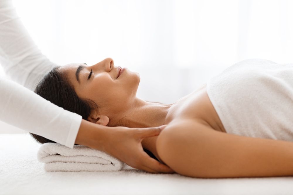 Woman receiving a neck massage, eyes closed, on a white towel, in a bright room.