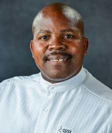 A person smiling, wearing a light-colored, long-sleeved shirt with a Mandarin collar, against a plain gray background.