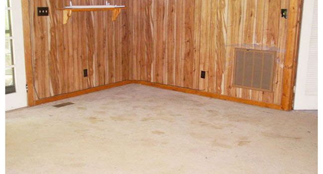 An empty corner room with vertical wood paneling, carpeted floors, a wall vent, and a small wooden shelf.