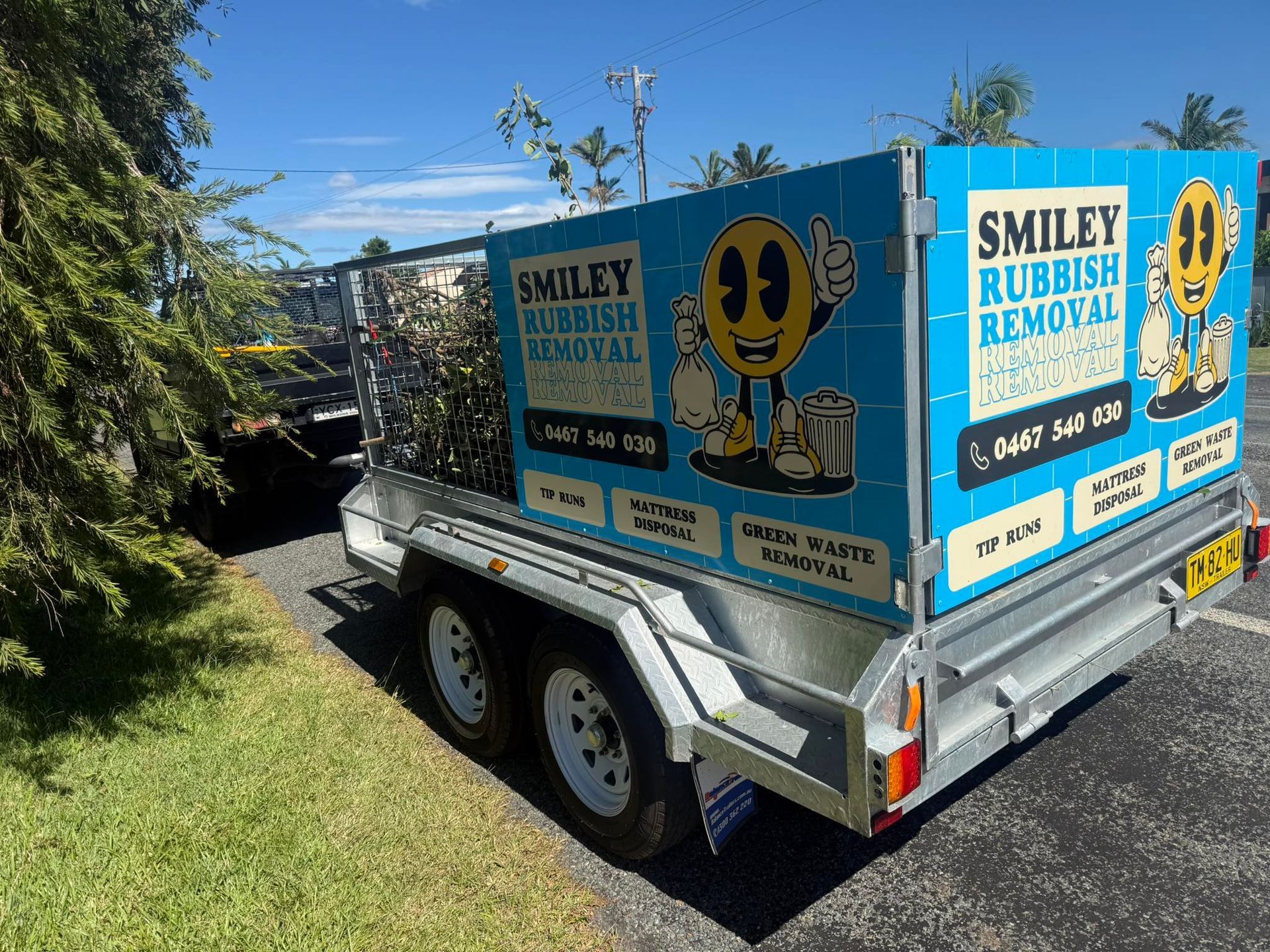 A trailer for Smiley Rubbish Removal featuring a yellow smiley face logo on a blue background, parked near green trees.