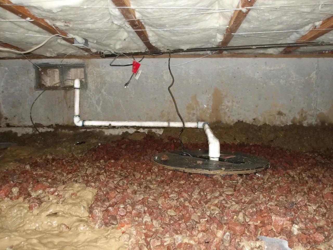 Basement with sump pump and gravel ground. White pipes, exposed beams, and concrete wall. Muddy areas present.