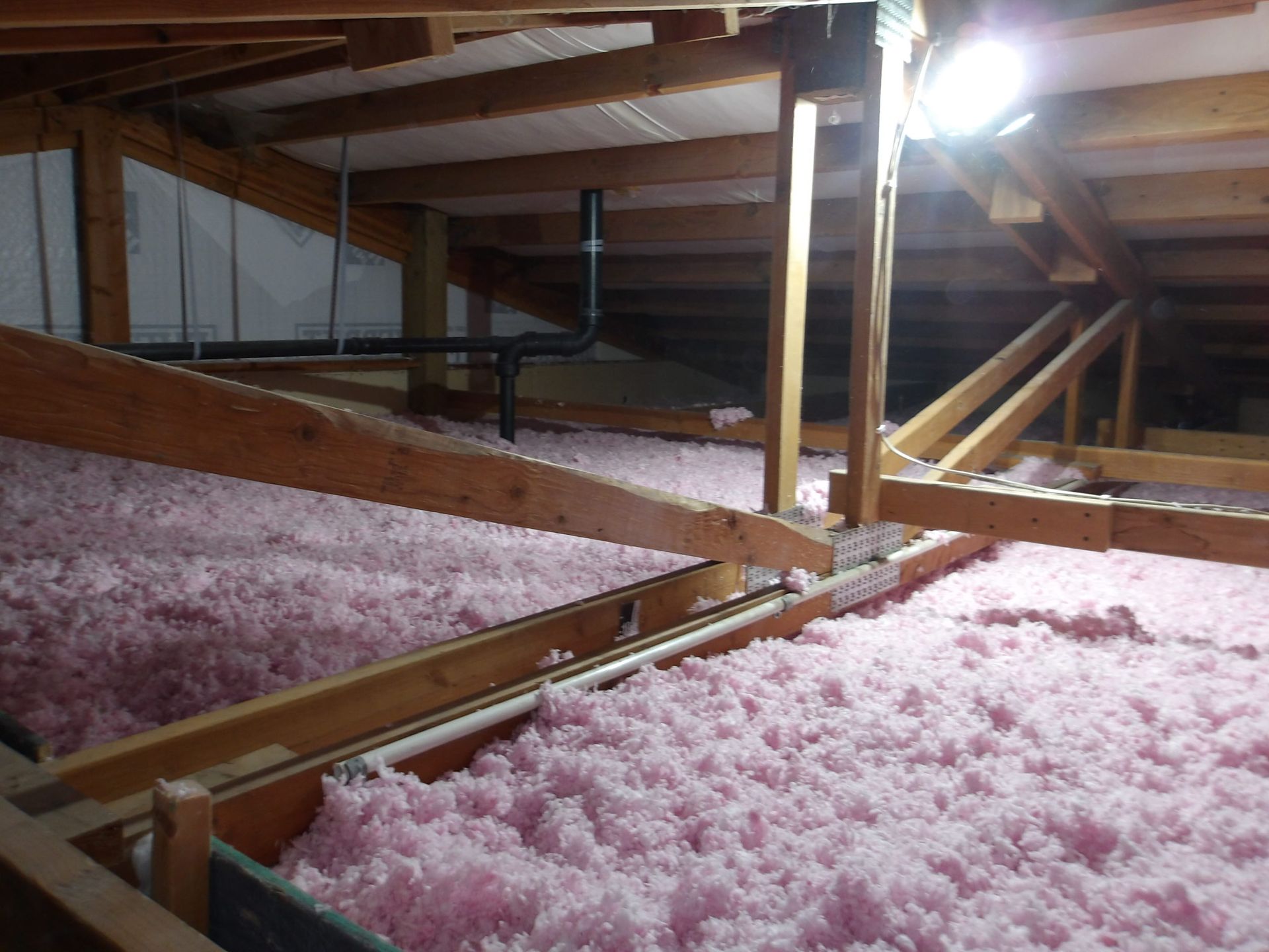 Attic interior featuring light pink blown-in insulation between wooden ceiling joists and a black plumbing vent pipe.