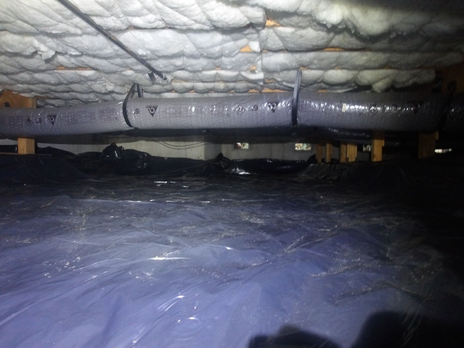 A view of a crawlspace with a blue vapor barrier on the ground, insulated floor joists, and suspended air ducts.