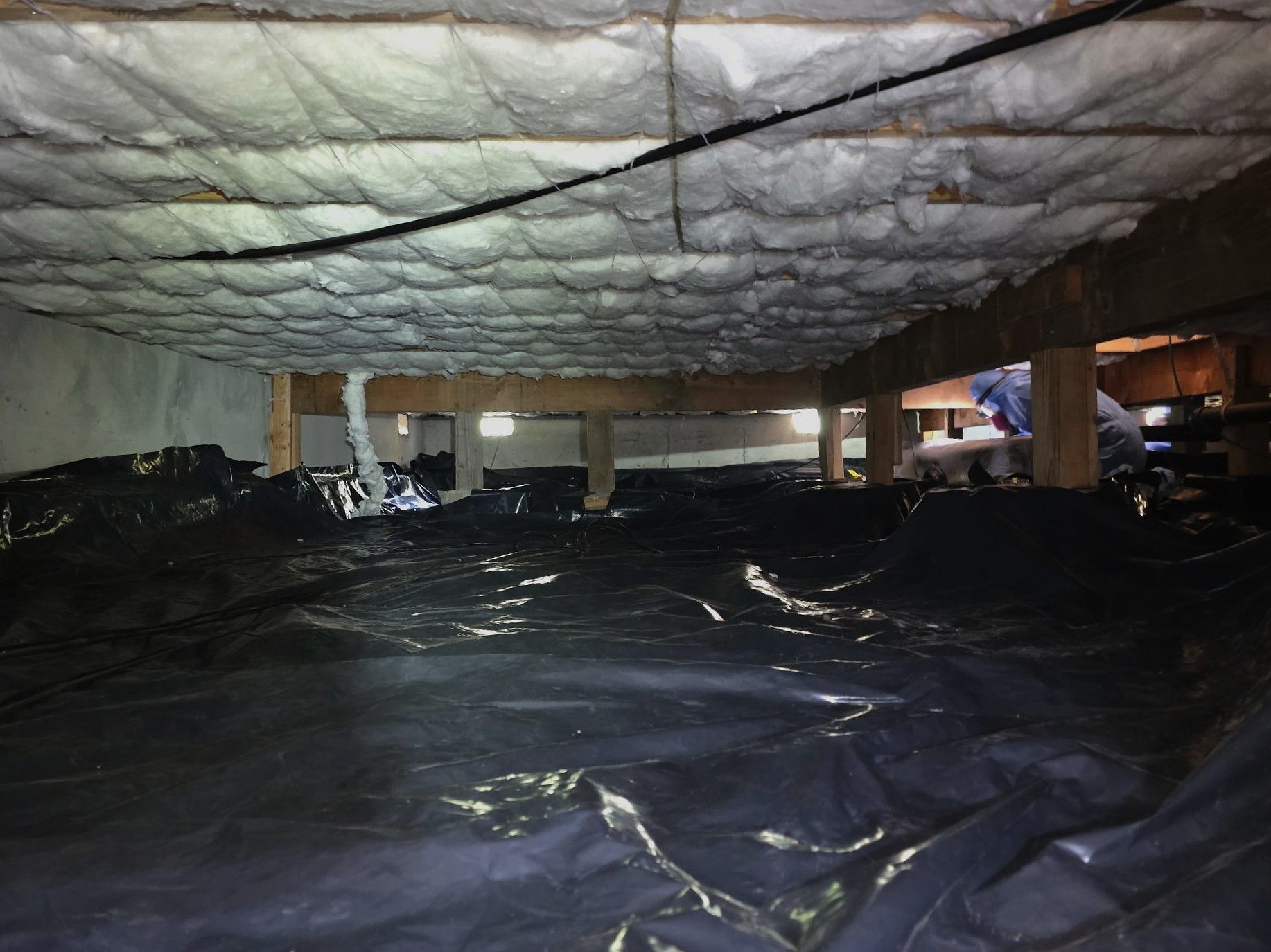 A crawl space with a dark plastic ground vapor barrier and white spray foam insulation between the wooden floor joists.