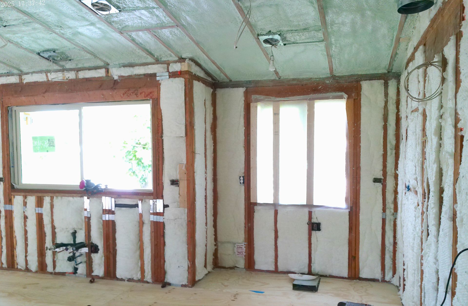 Interior room under construction featuring wooden wall studs, exposed window frames, and white spray foam insulation.