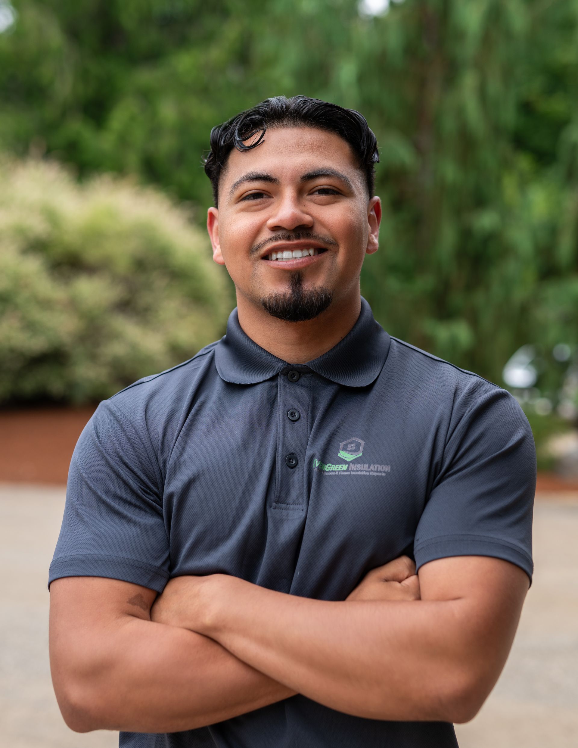 Man in grey polo shirt, arms crossed, smiling outdoors.