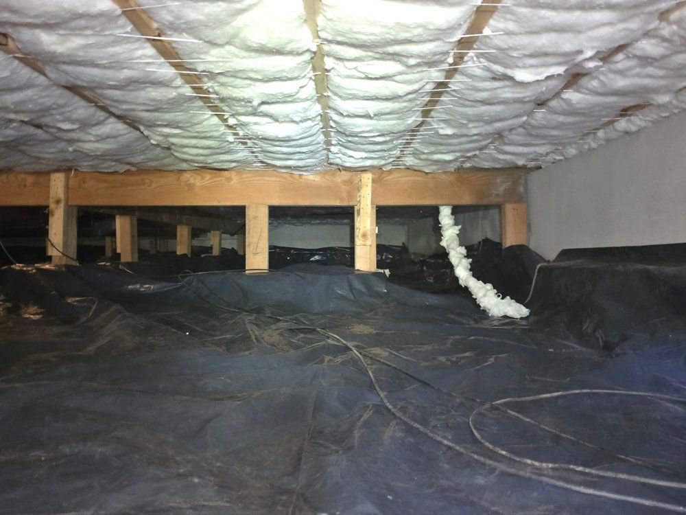 Crawl space with wooden beams, insulation, and black vapor barrier on the ground.