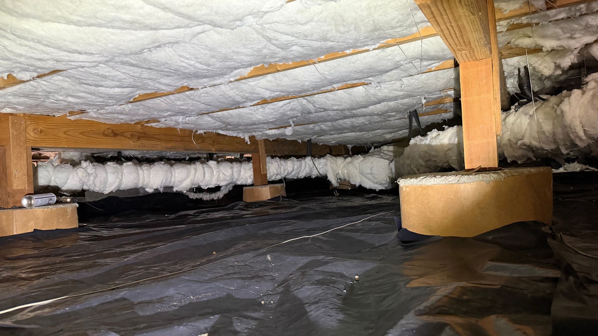 A crawl space with foam insulation on the floor joists, a vapor barrier on the ground, and supporting wooden piers.