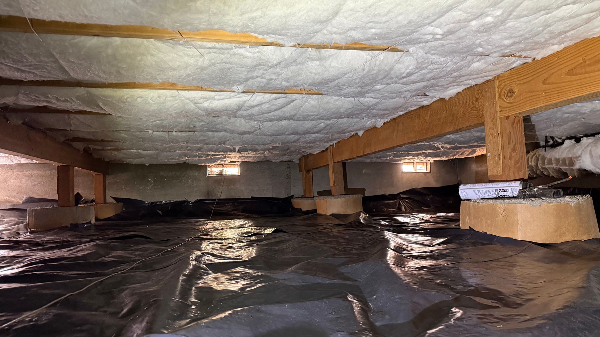 A crawl space with foam insulation sprayed on the ceiling joists, a black vapor barrier on the ground, and concrete piers.