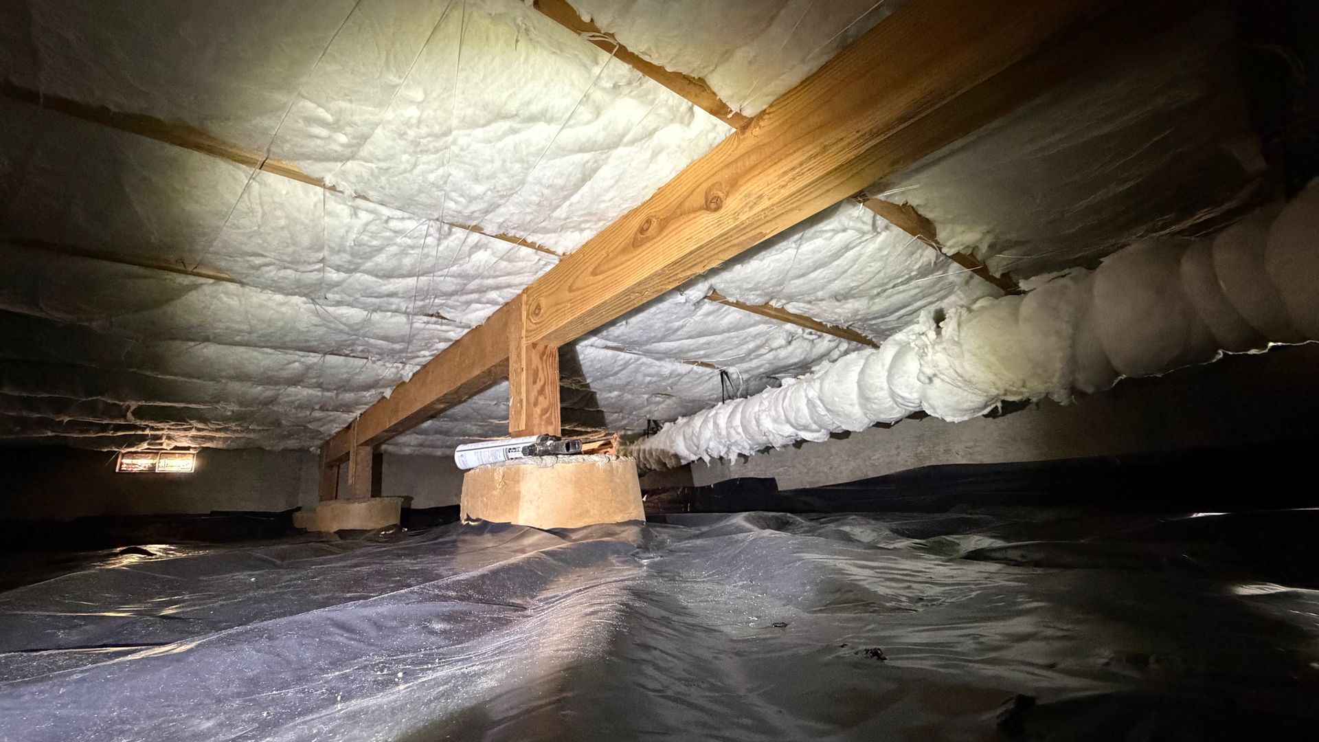 A crawl space with foam insulation on the subfloor, wooden supports, and a plastic vapor barrier covering the ground.