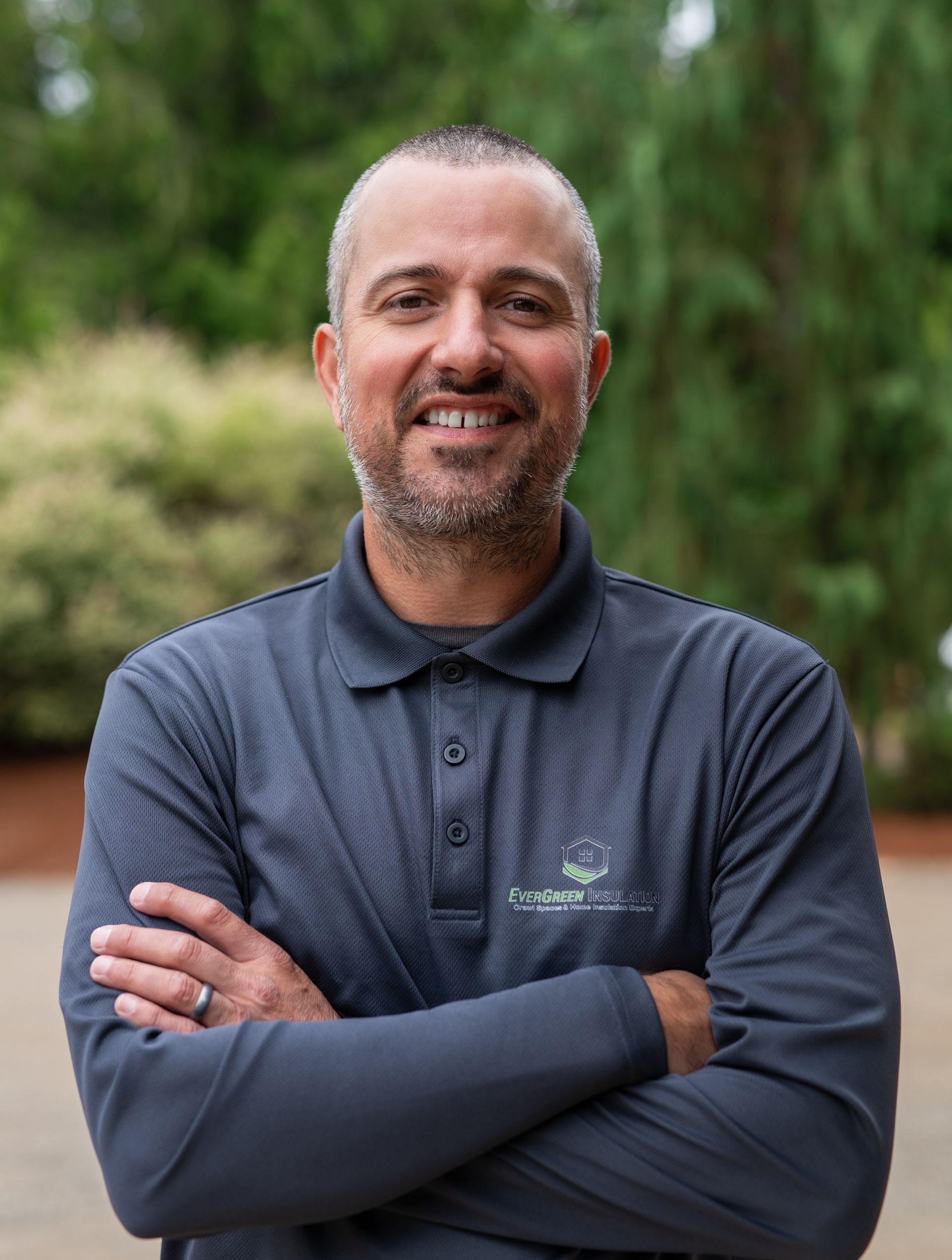 Man with arms crossed, wearing a gray polo shirt, smiling, set outdoors in front of greenery.
