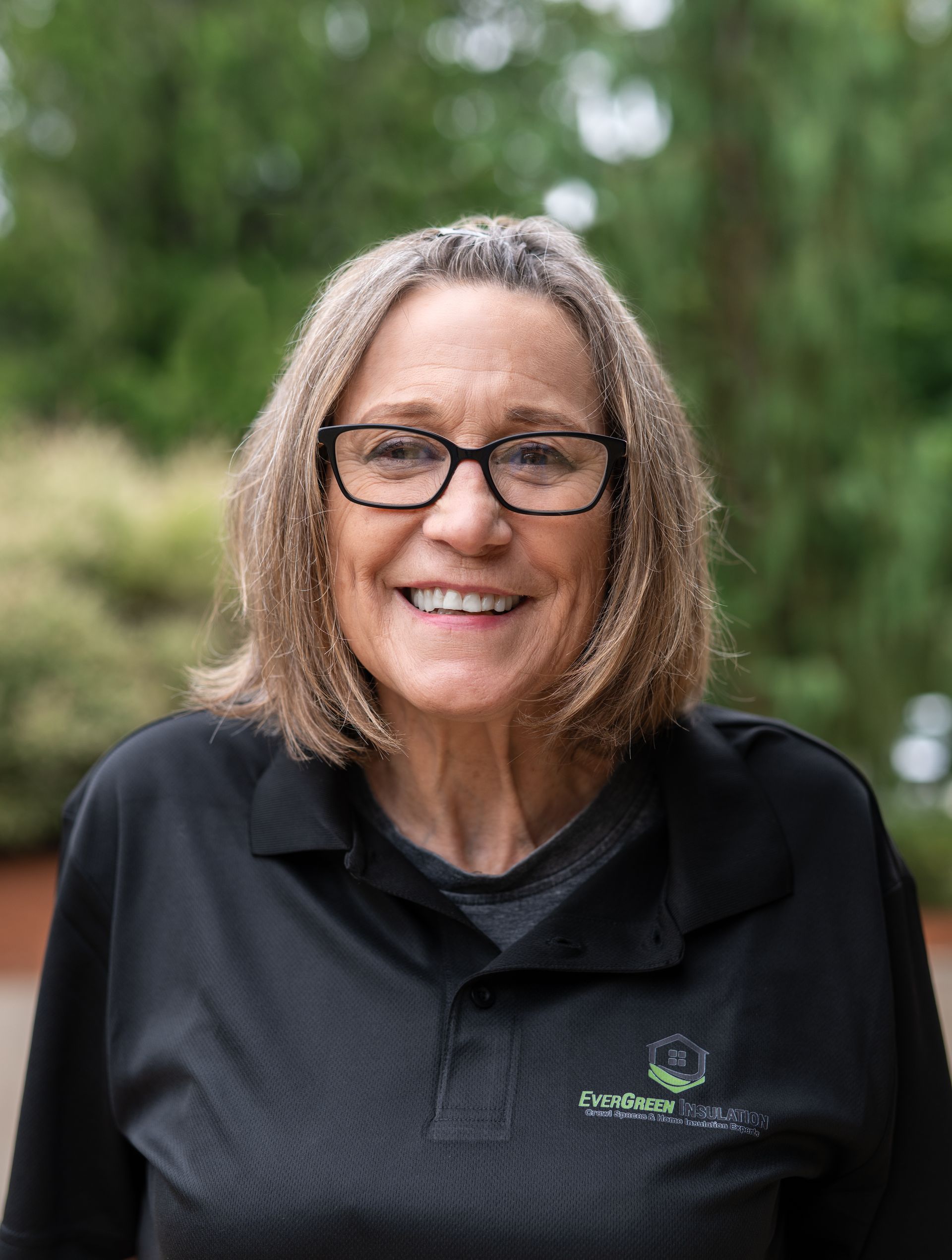 Woman wearing glasses, smiling, in a black shirt with logo, outdoors with greenery in background.