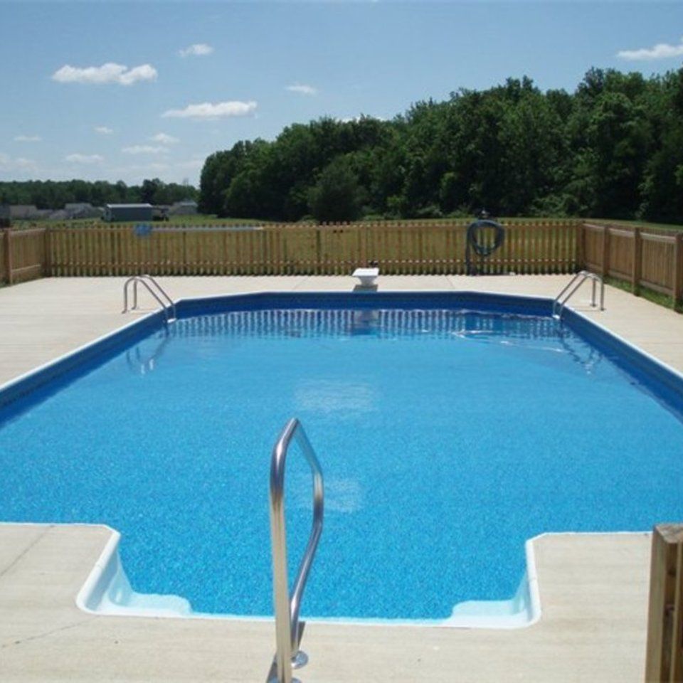 Outdoor swimming pool with blue water, white deck, and metal handrails, surrounded by a wooden fence and trees.