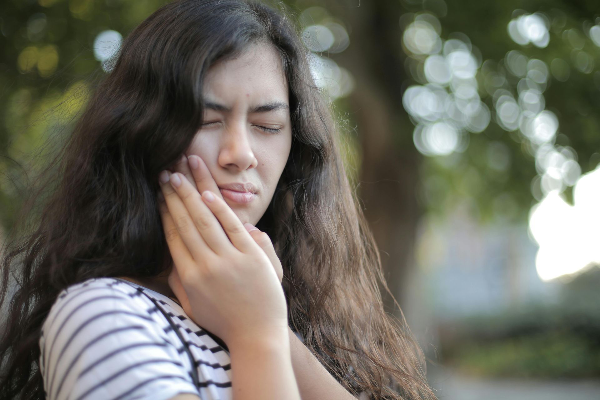 Woman with long brown hair, eyes closed, touching her cheek, possibly in pain, outdoors.