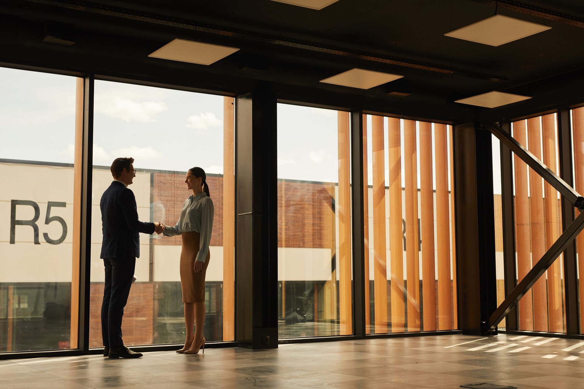 Two people shaking hands in a large modern room with floor-to-ceiling windows.