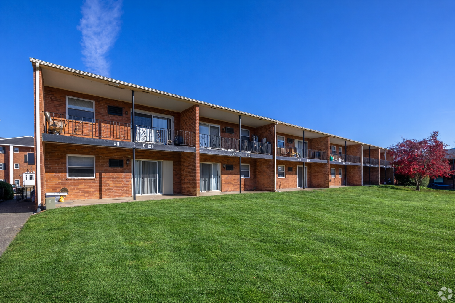 A two-story brick apartment building with balconies, set against a clear blue sky above a vibrant green lawn.