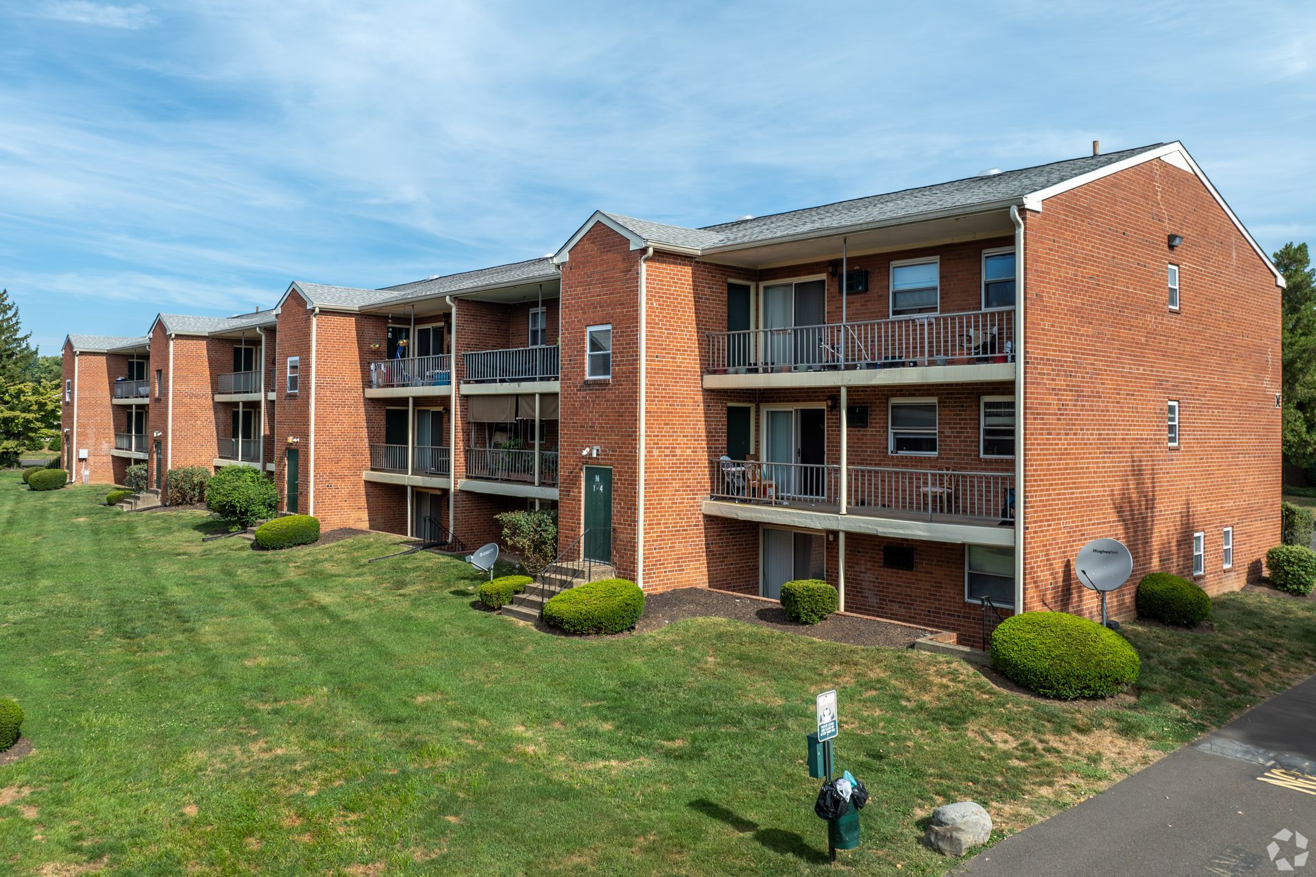 A three-story red brick apartment building with recessed balconies and a green lawn under a blue, cloudy sky.