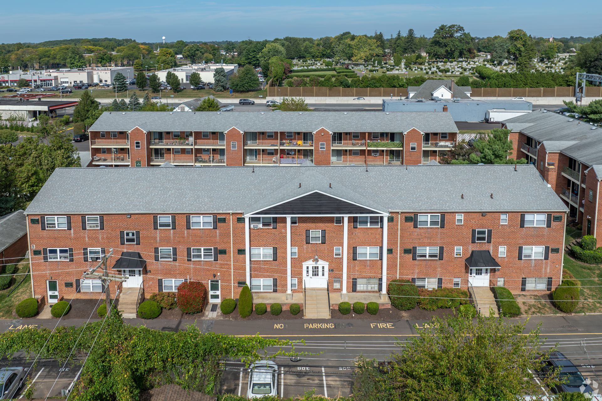 An aerial view of a three-story brick apartment complex with a gray shingled roof and a parking lot in the foreground.