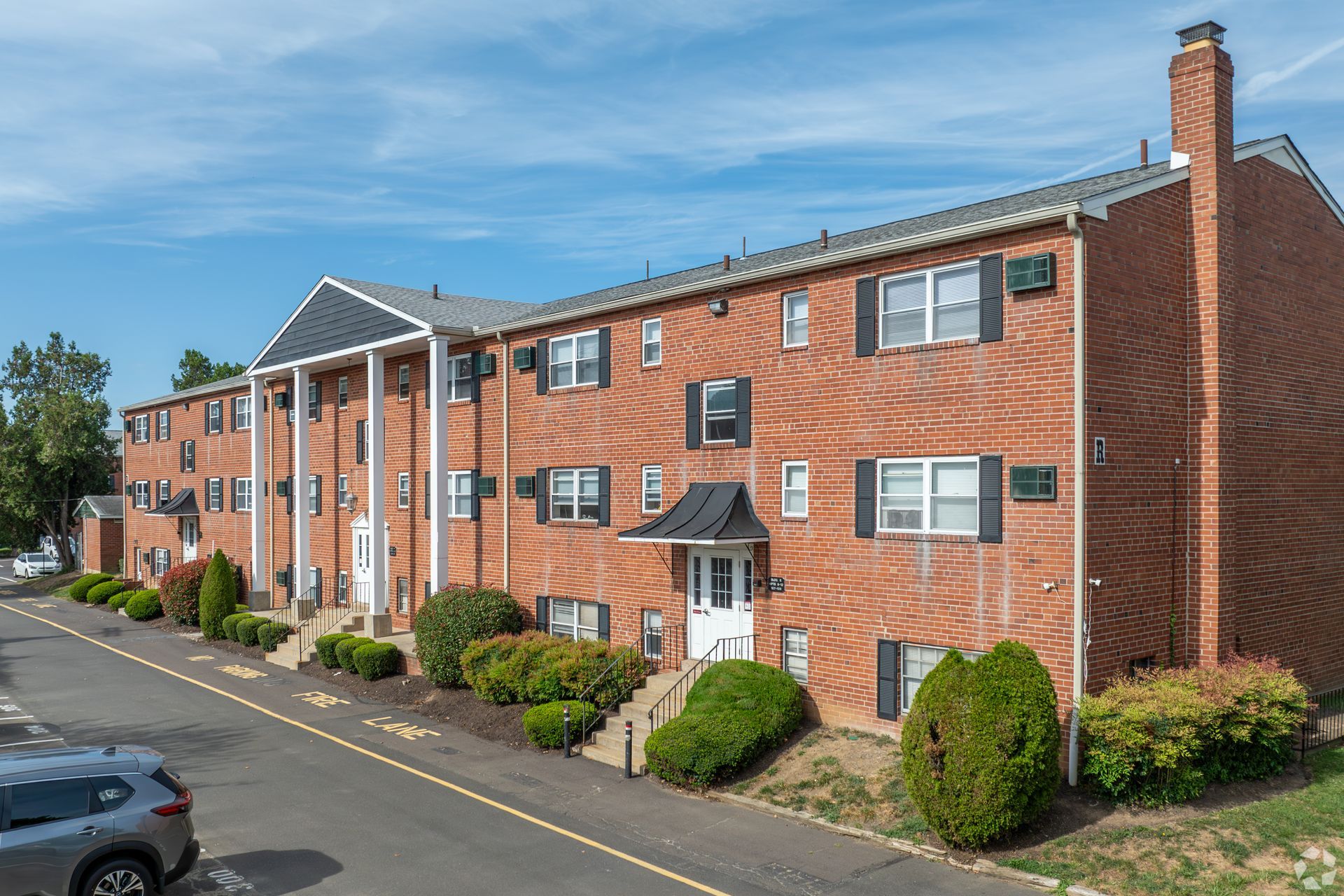 Three-story red brick apartment building with white columns at the entrance, a chimney, and a small parking lot.