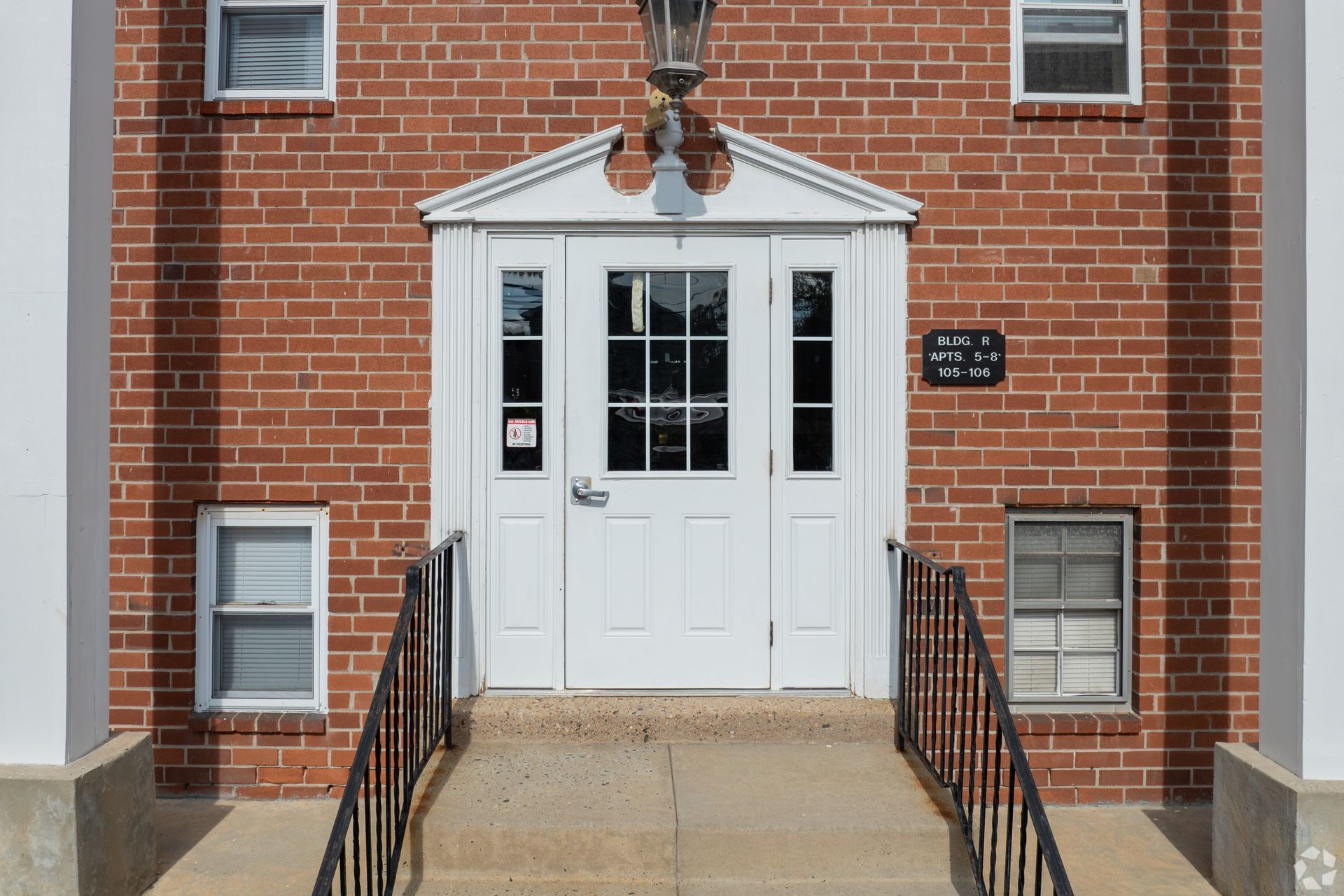 A brick building entrance with a white door, small window panes, a black metal railing, and a triangular architectural hood.