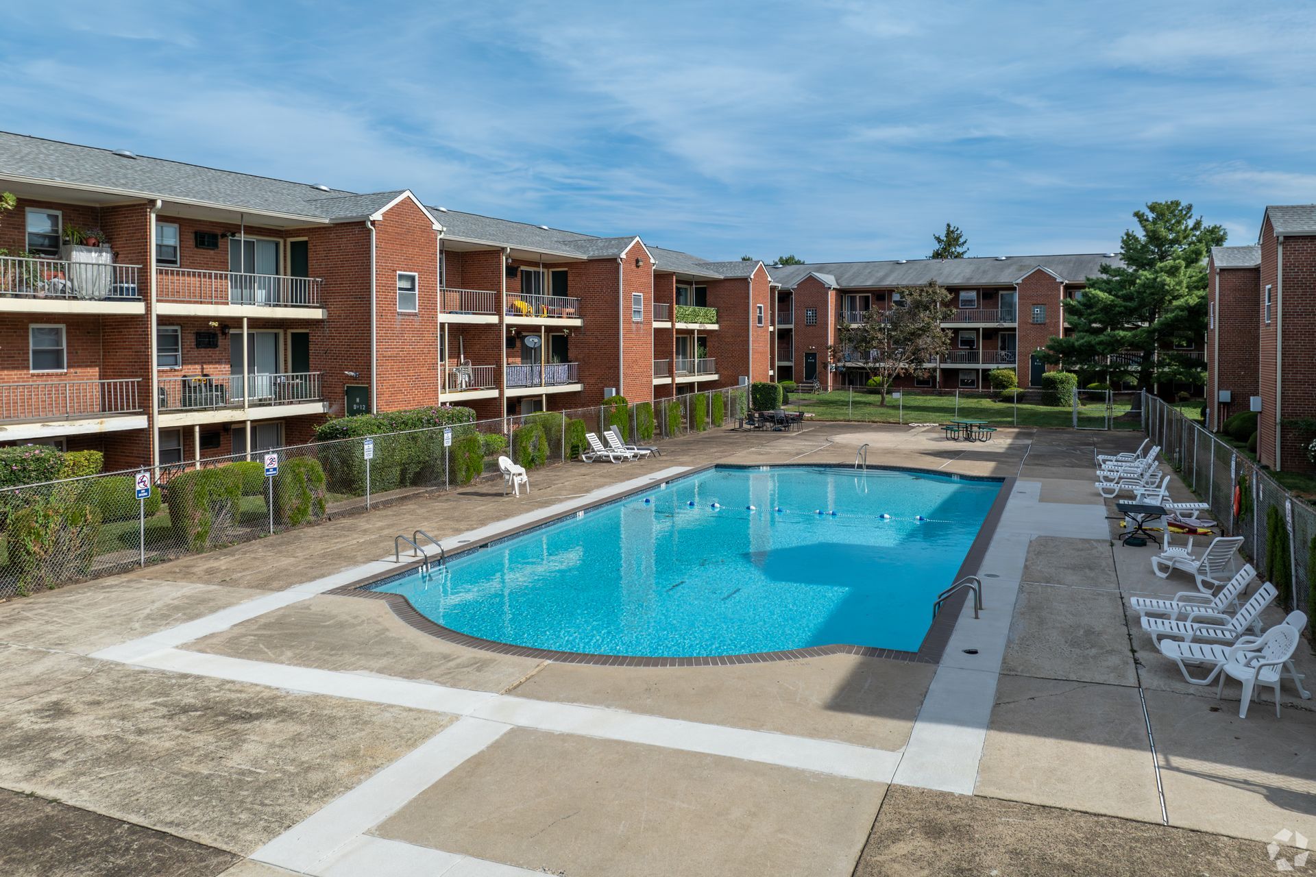 A rectangular swimming pool surrounded by a concrete deck and a multi-story brick apartment building under a blue sky.