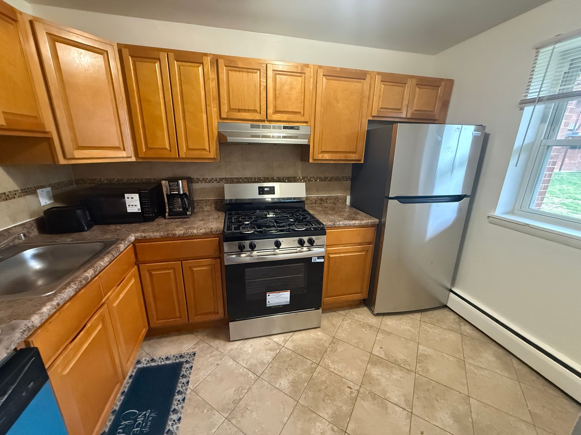 A kitchen with honey-oak cabinets, stainless steel appliances, a tiled floor, and a window to the right.