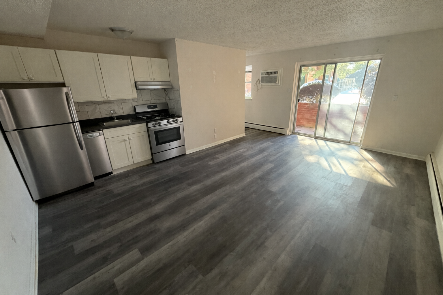 An open-concept apartment with a kitchen featuring stainless steel appliances, white cabinets, and gray wood-look flooring.