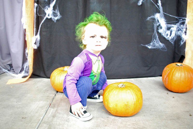 A little boy dressed as the joker is squatting next to pumpkins