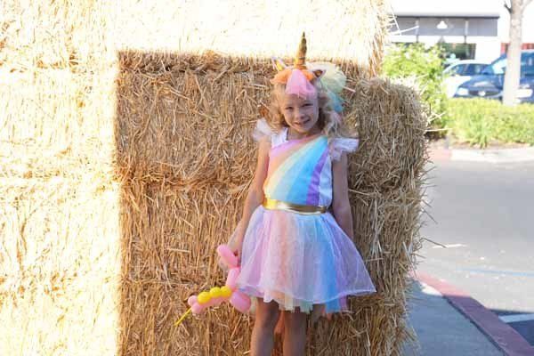 A little girl in a unicorn costume is standing next to a bale of hay.